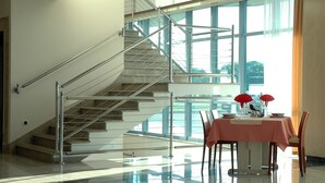 Un hall d’hôtel moderne avec un escalier, un coin repas avec une table dressée pour deux personnes, et de grandes fenêtres offrant une vue sur l’extérieur.