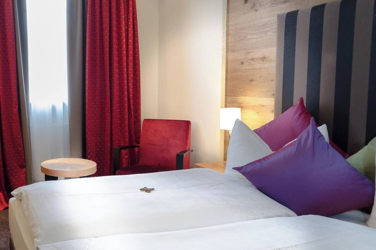 Quarto de hotel com cama, cortinas vermelhas, poltrona vermelha, mesa redonda pequena e uma luminária.