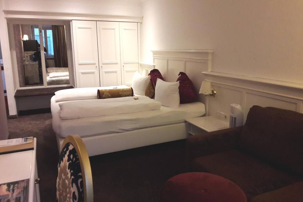 Quarto de hotel com cama de casal, uma mesinha de cabeceira, um banquinho vermelho e uma janela com cortinas.