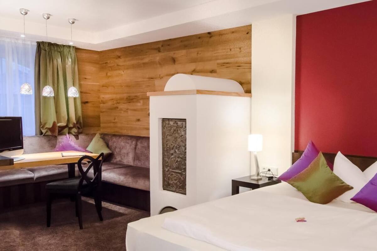 Quarto de hotel com parede de destaque em madeira, cama com almofadas coloridas, escrivaninha com cadeira e um sofá.