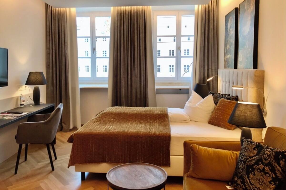 Quarto de hotel com cama, escrivaninha, cadeira, mesa pequena e sofá. Há cortinas nas janelas e um quadro na parede.