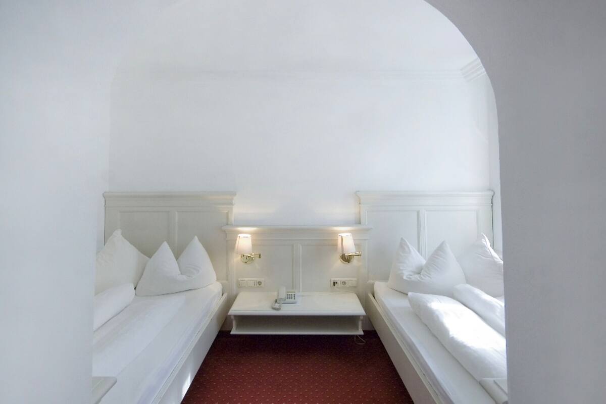Quarto de hotel com duas camas brancas, uma mesinha e duas luminárias fixadas na parede.