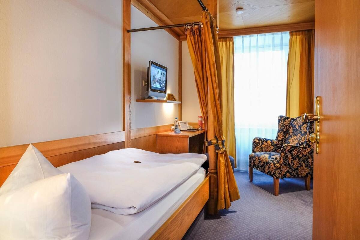 Quarto de hotel com cama, escrivaninha, cadeira e janela com cortinas.