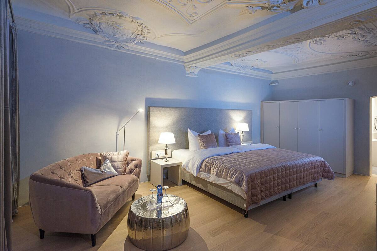Quarto com cama grande, sofá, mesa de cabeceira, guarda-roupa e detalhes ornamentais no teto.