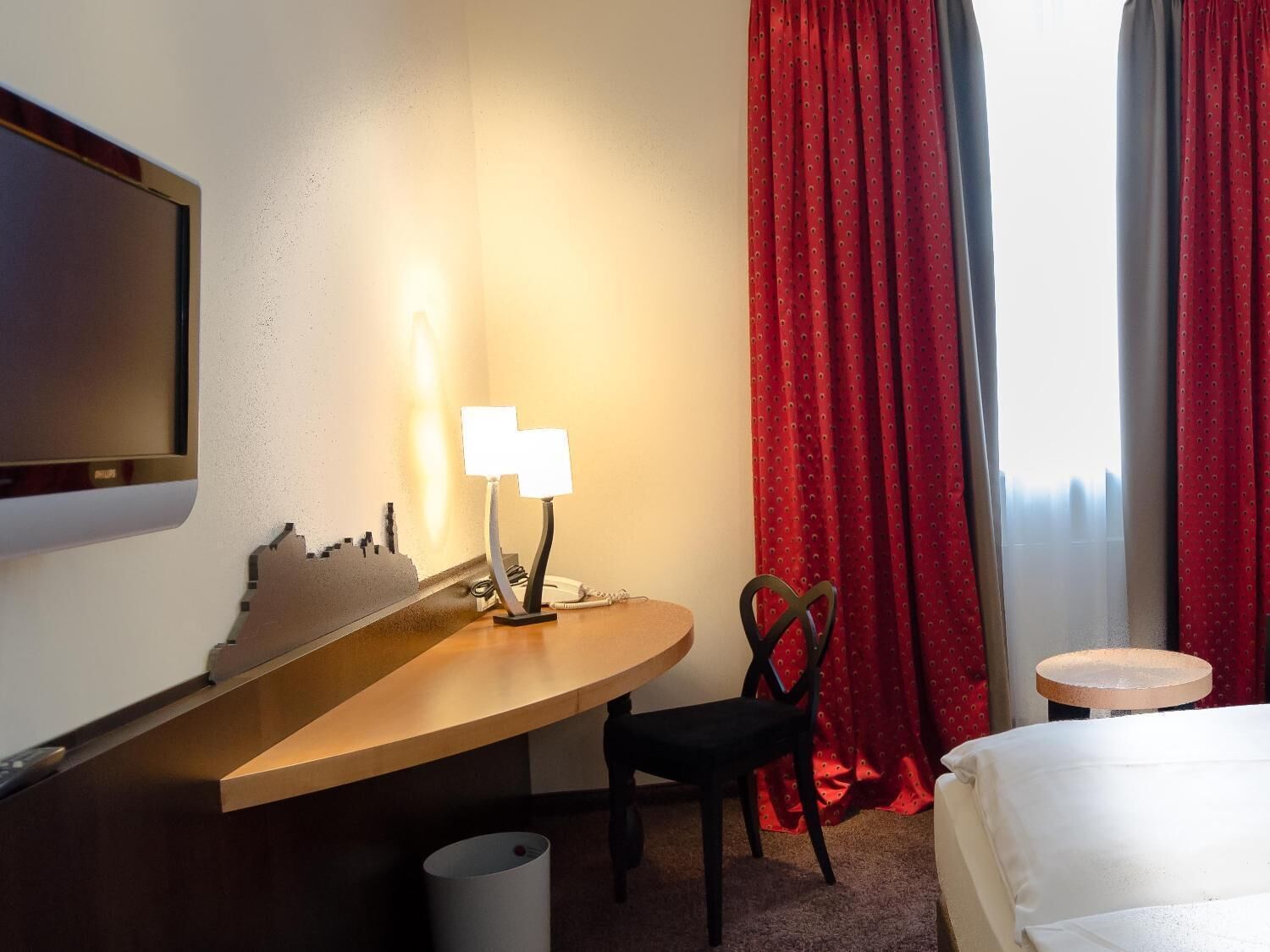 A hotel room with a bed, a desk with a lamp, a chair, a TV, and a window with red curtains.