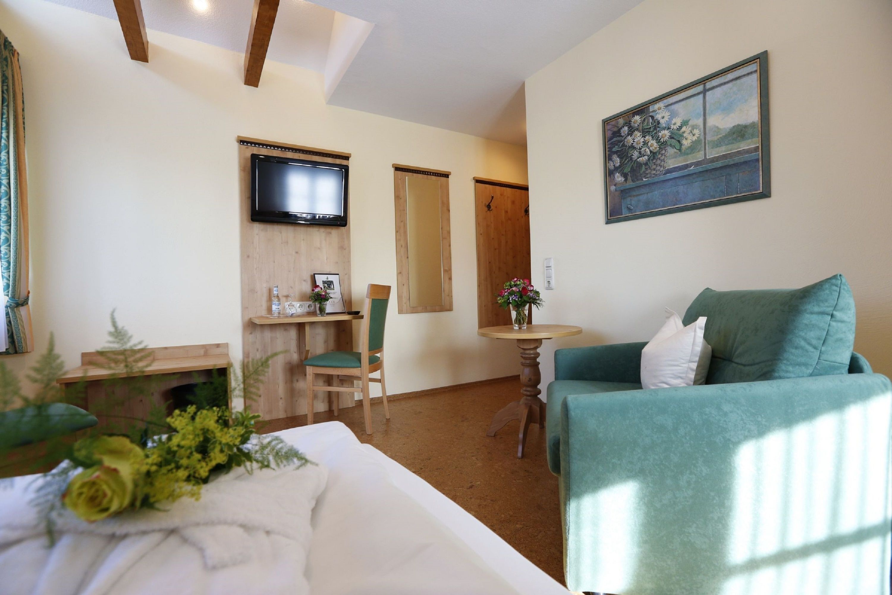 A hotel room with a bed, a green armchair, a small table with a vase of flowers, a television mounted on the wall, and a painting above the door.