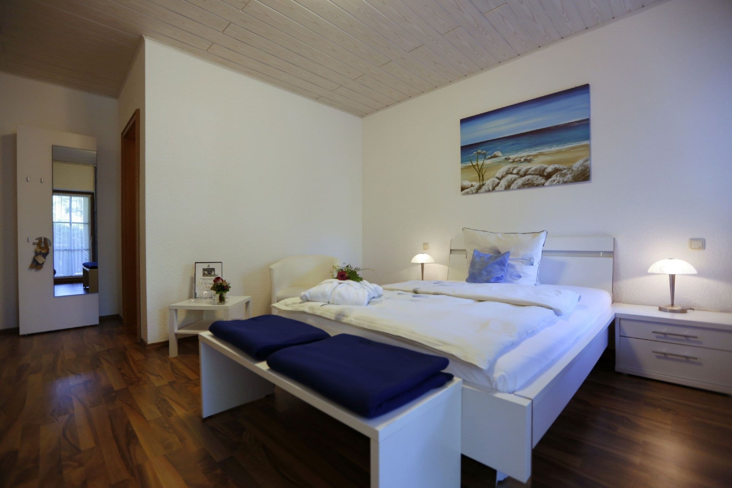 A bedroom with a bed, bedside tables, a bench, a painting, and a window.