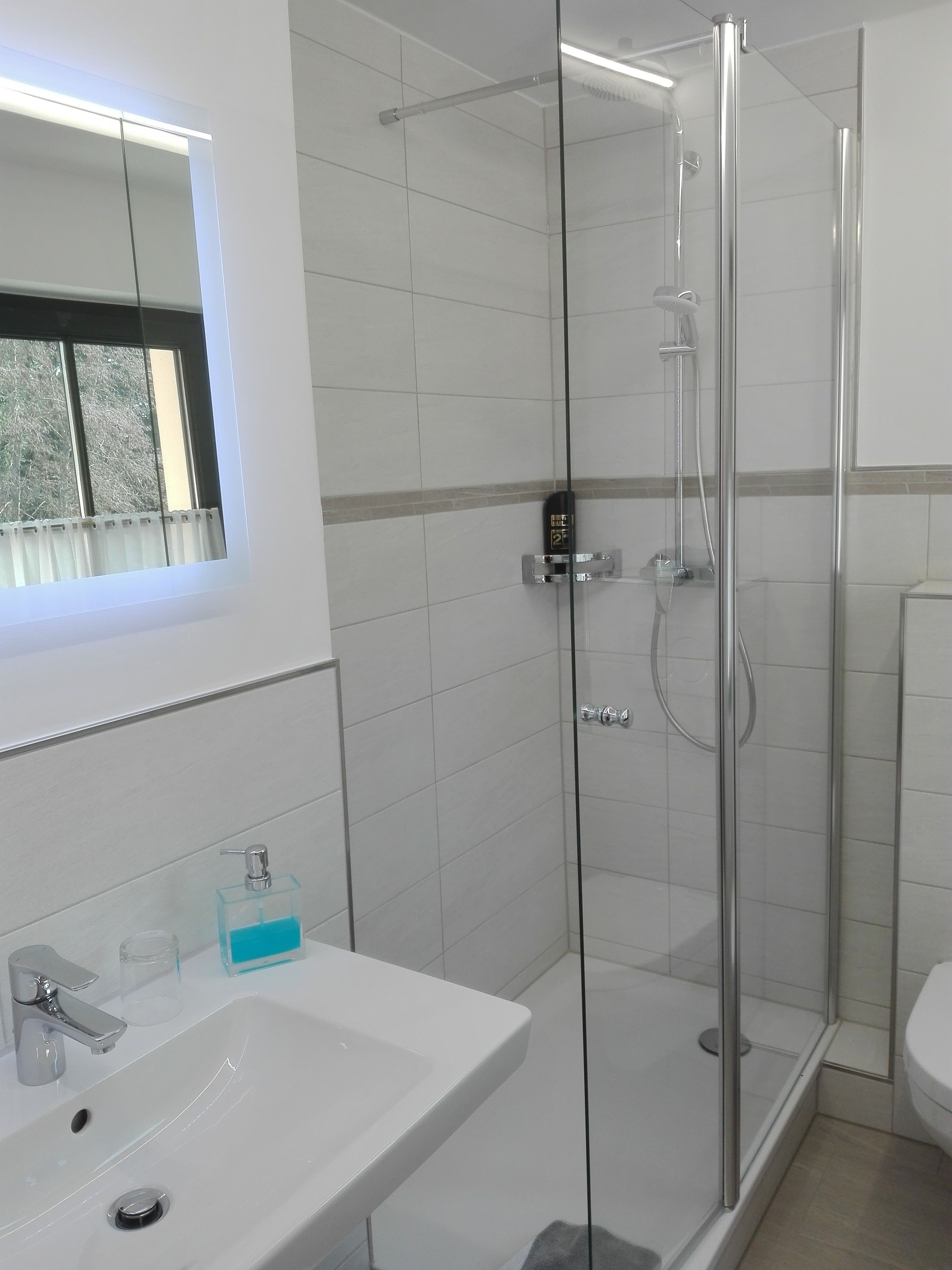 Een moderne badkamer met een douchecabine van glas, een witte wastafel met een chromen kraan en een raam boven de wastafel.