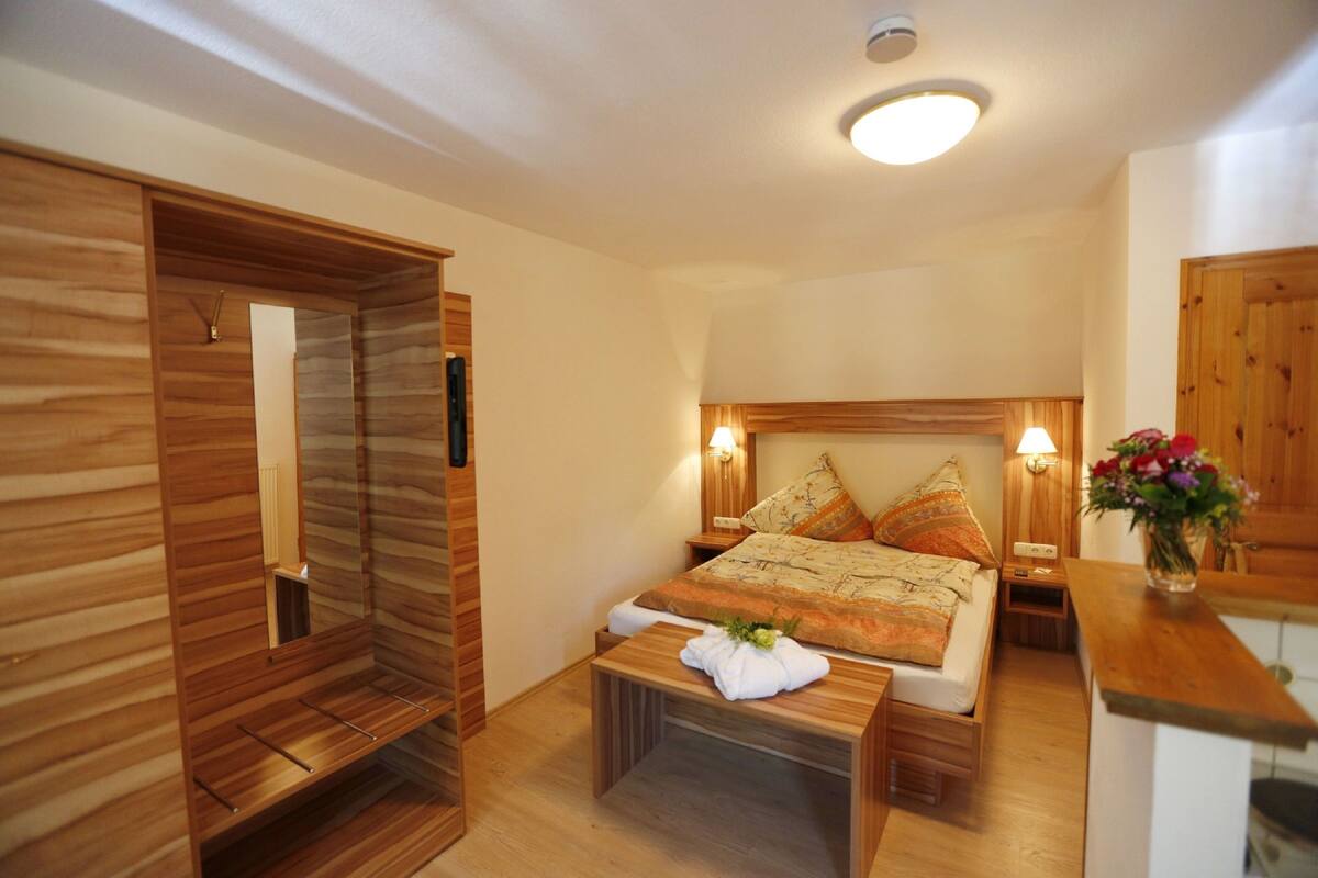 A bedroom with a wooden wardrobe, a bed with patterned bedding, bedside lamps, and a small table with a towel.