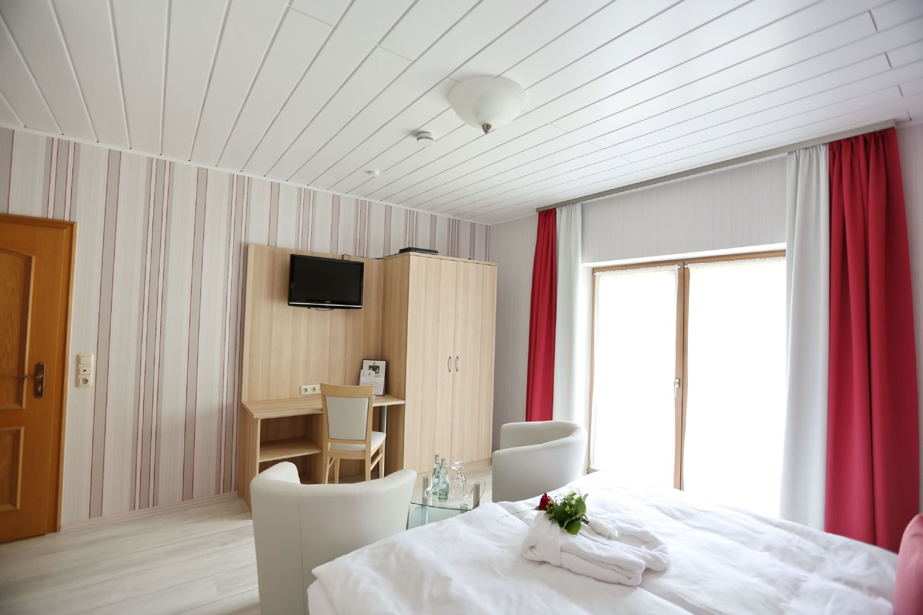 A hotel room with a bed, a desk, a chair, a wardrobe, a television, and a window with curtains.