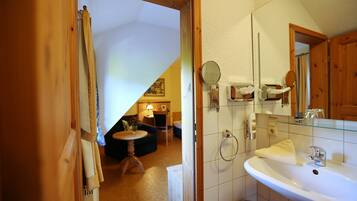 Un baño con lavamanos blanco, espejo y perchero. Hay una puerta de madera que da a una sala de estar con sofá y lámpara.