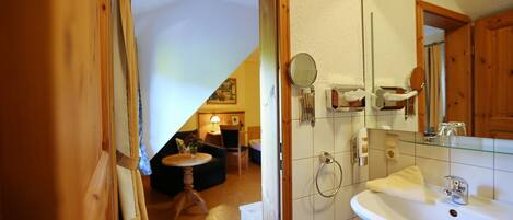 Ein Badezimmer mit weißem Waschbecken, Spiegel und Handtuchhalter. Eine Holztür führt in einen Wohnbereich mit Sofa und Lampe.