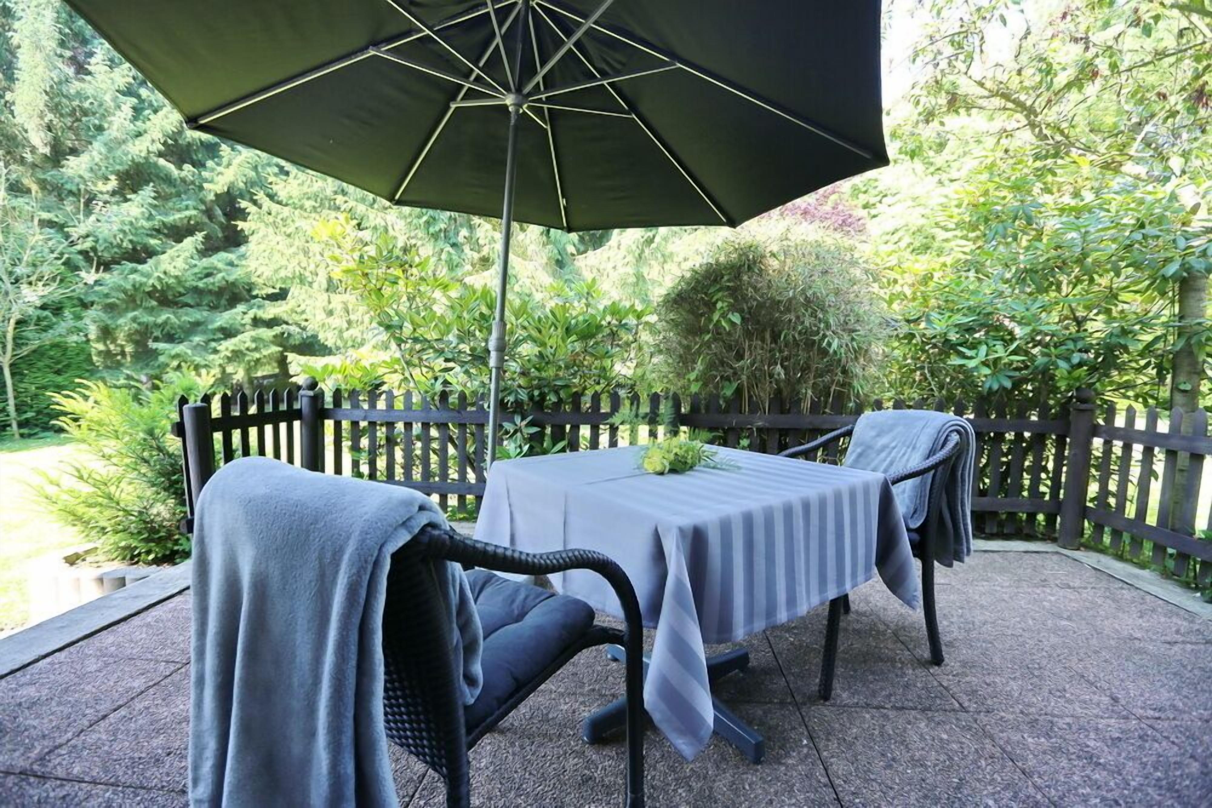 Eine Terrassenanlage im Freien mit zwei Tischen, die mit grauen Tischdecken bedeckt sind, einem großen Sonnenschirm und einem schwarzen Zaun, der den Bereich umgibt.
