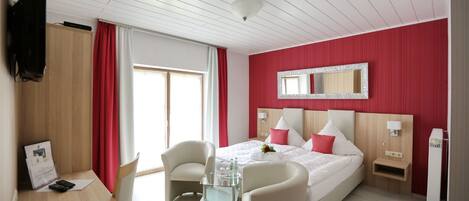 Habitación de hotel con cama, dos sillas, una mesita, televisor, espejo y ventana con cortinas.