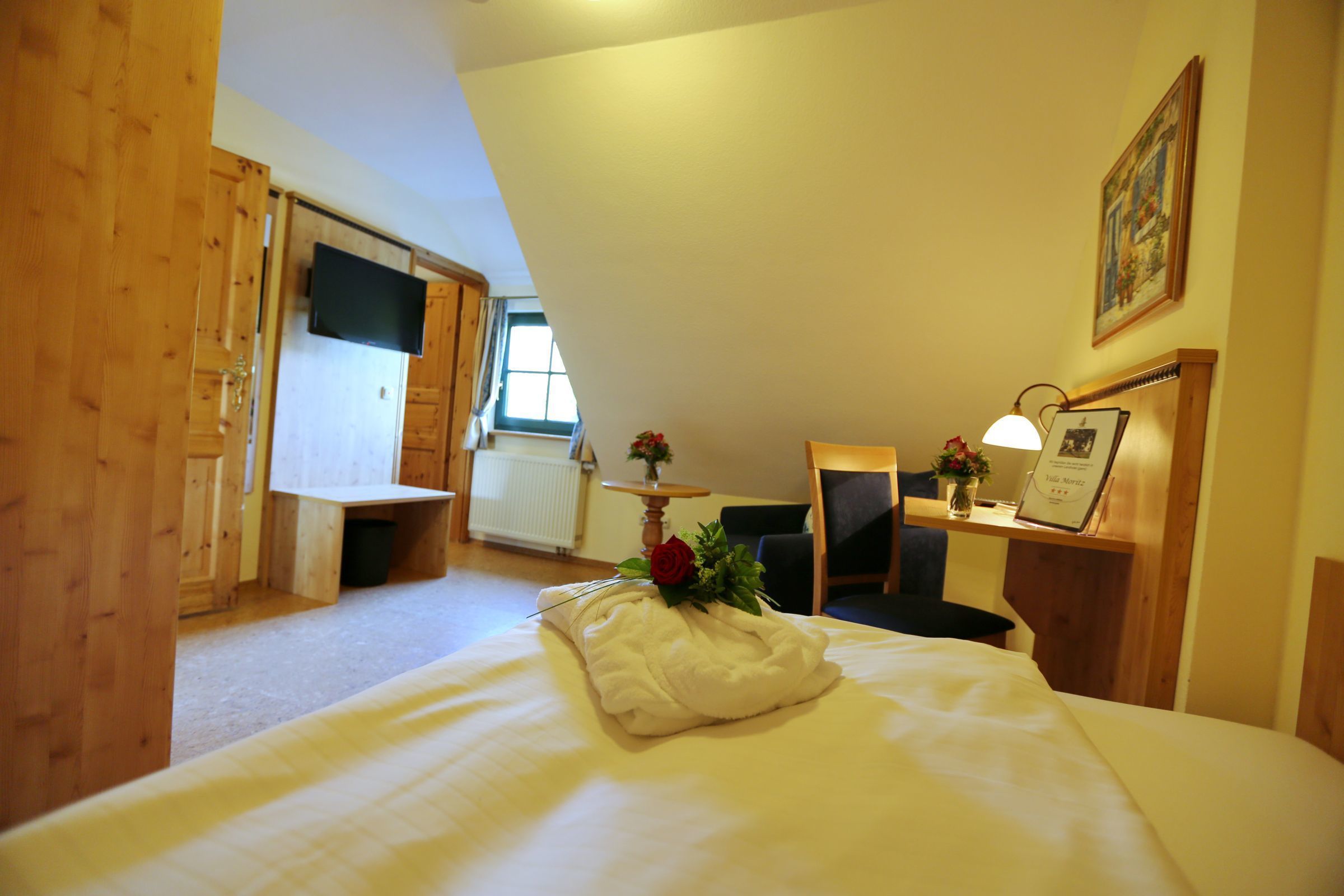 A bedroom with a bed, a desk, a chair, a small table with a vase and a picture frame, and a window.