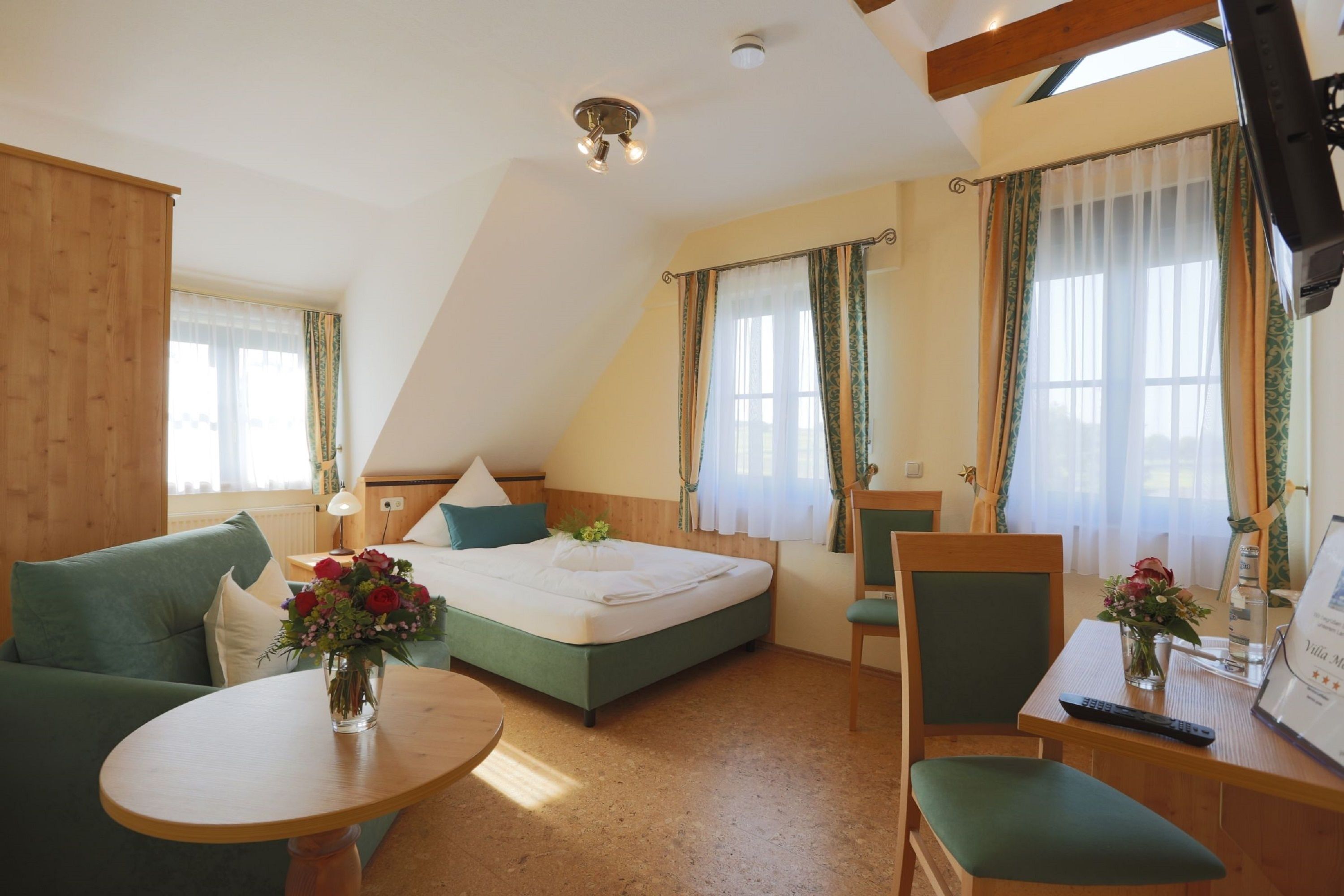 A bedroom with a bed, a green sofa, a small table with a vase of flowers, and a TV on a wooden stand.