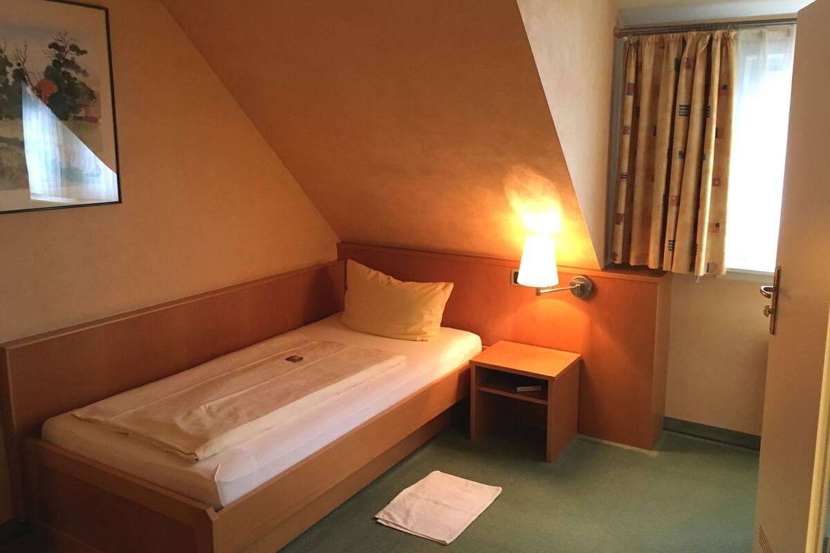 A small, single-bed room with a wooden headboard, a bedside table, a wall-mounted lamp, and a window with curtains.