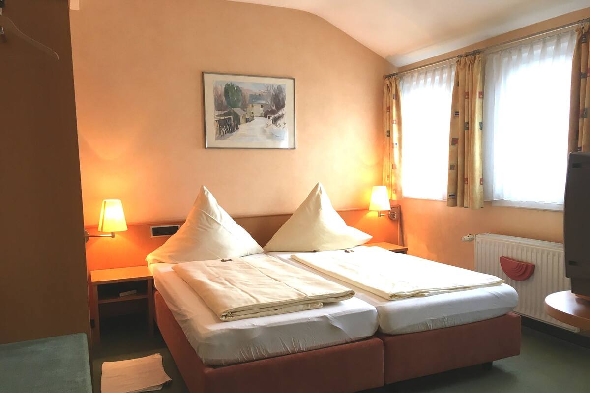 A hotel room with a bed, two bedside lamps, a painting on the wall, and a window with curtains.