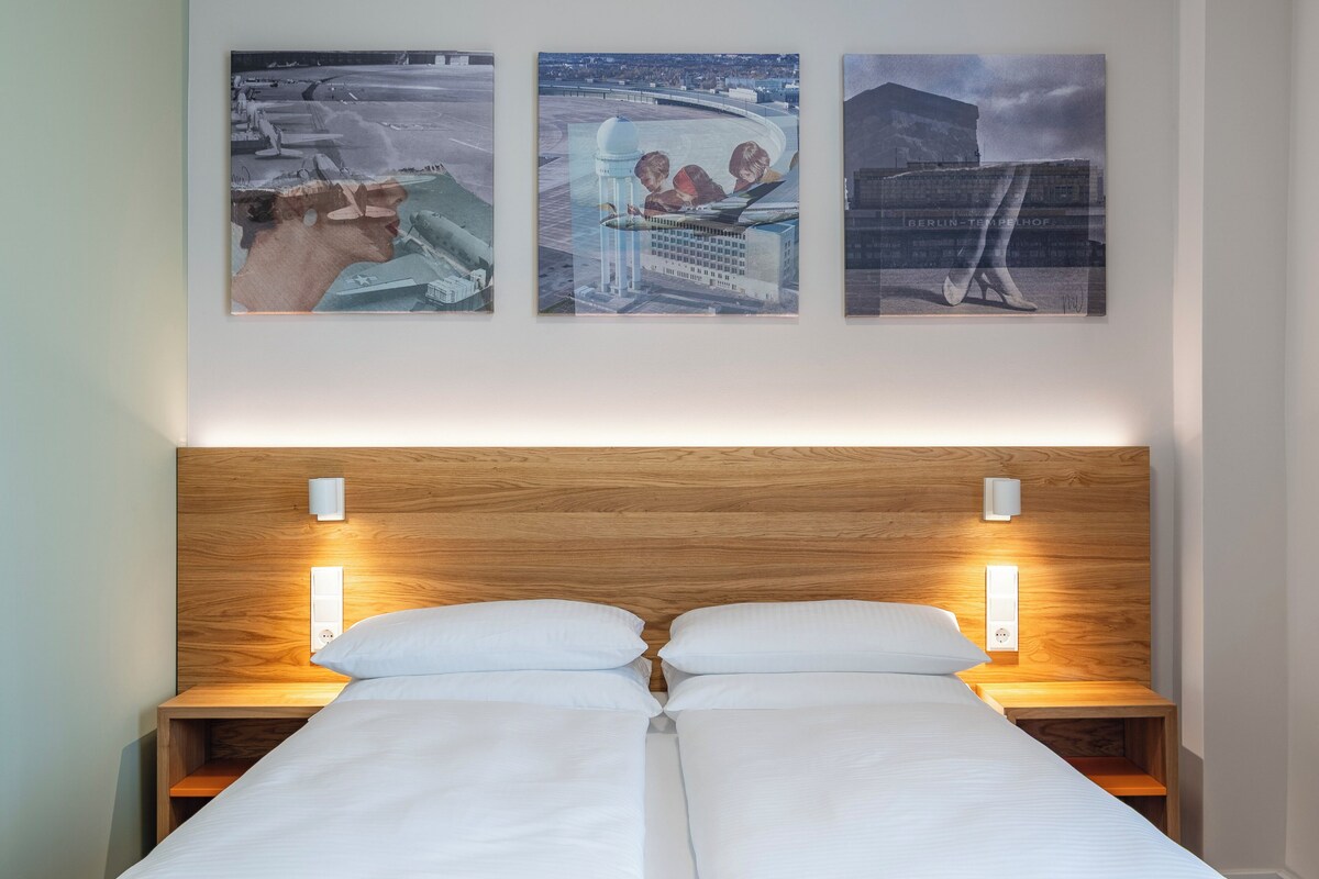 A hotel room with a wooden headboard, two bedside tables with lamps, a bed with white linens, and three framed artworks on the wall.