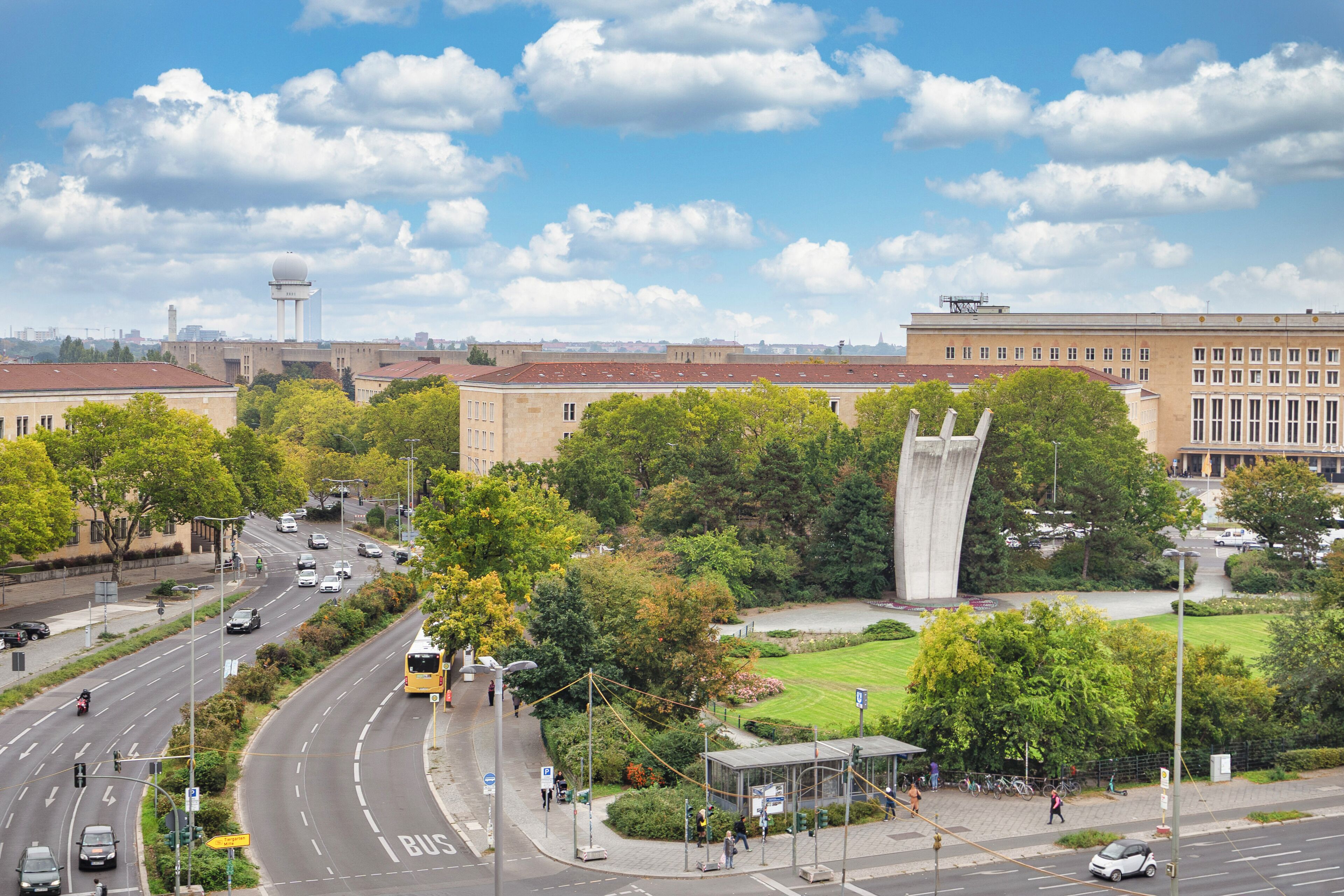 Eine Stadtansicht mit einem markanten Turm, einer Bushaltestelle und einer Mischung aus Grünflächen und Stadtgebäuden.