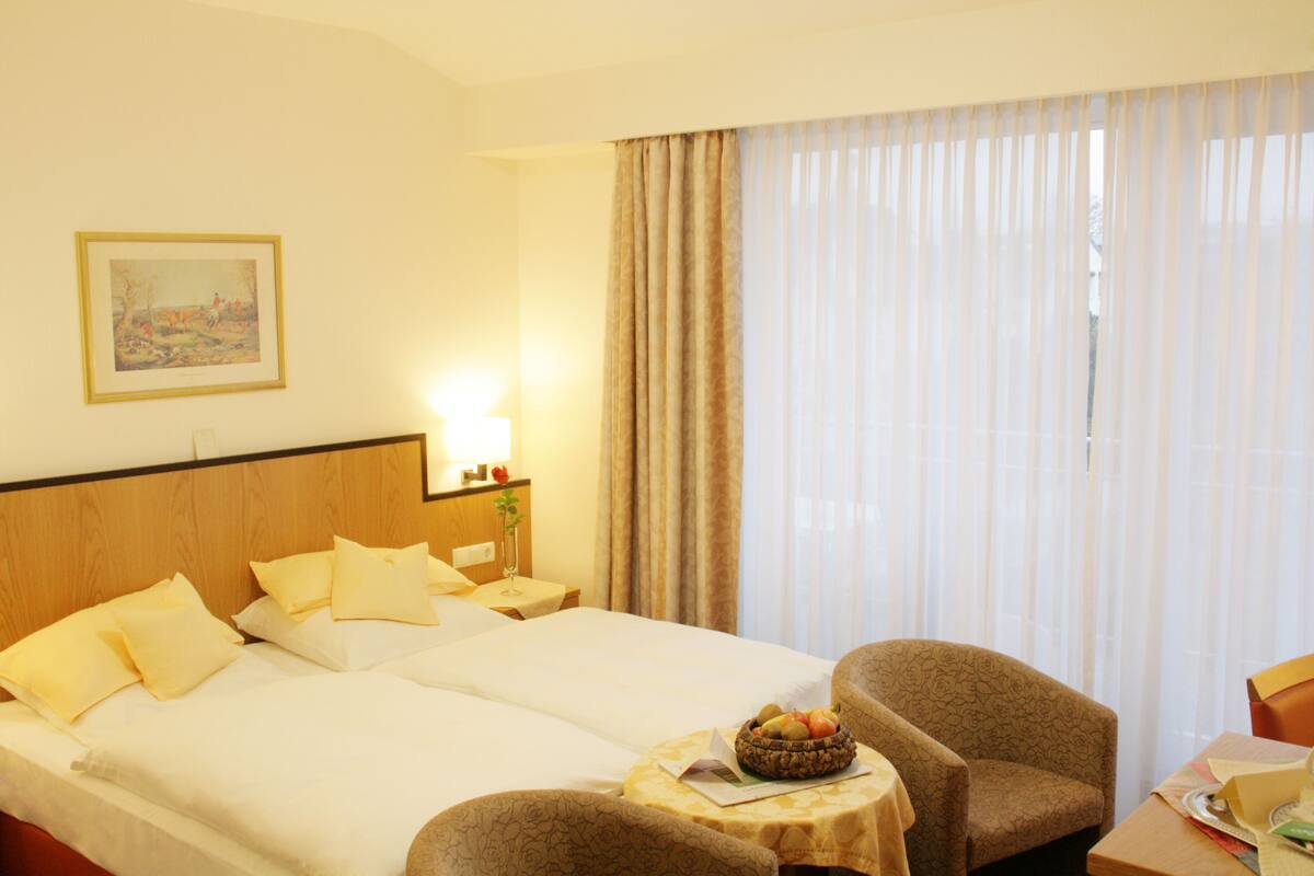 A hotel room with two beds, a small table with a fruit basket, a chair, a lamp, and a painting on the wall.