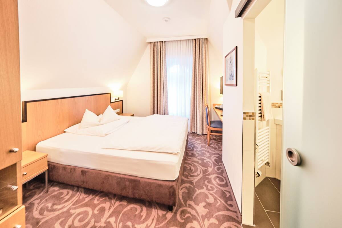 A hotel room with a bed, a nightstand, a desk, and a bathroom visible through an open door.