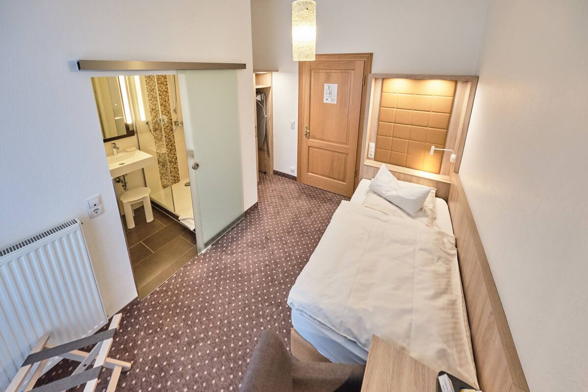 A compact hotel room with a bed, a small desk, a chair, a bathroom with a shower, and a toilet.