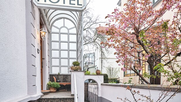 Entrada de un hotel con un amplio ventanal arqueado, un letrero que dice "HOTEL" y un pequeño jardín con plantas en macetas.