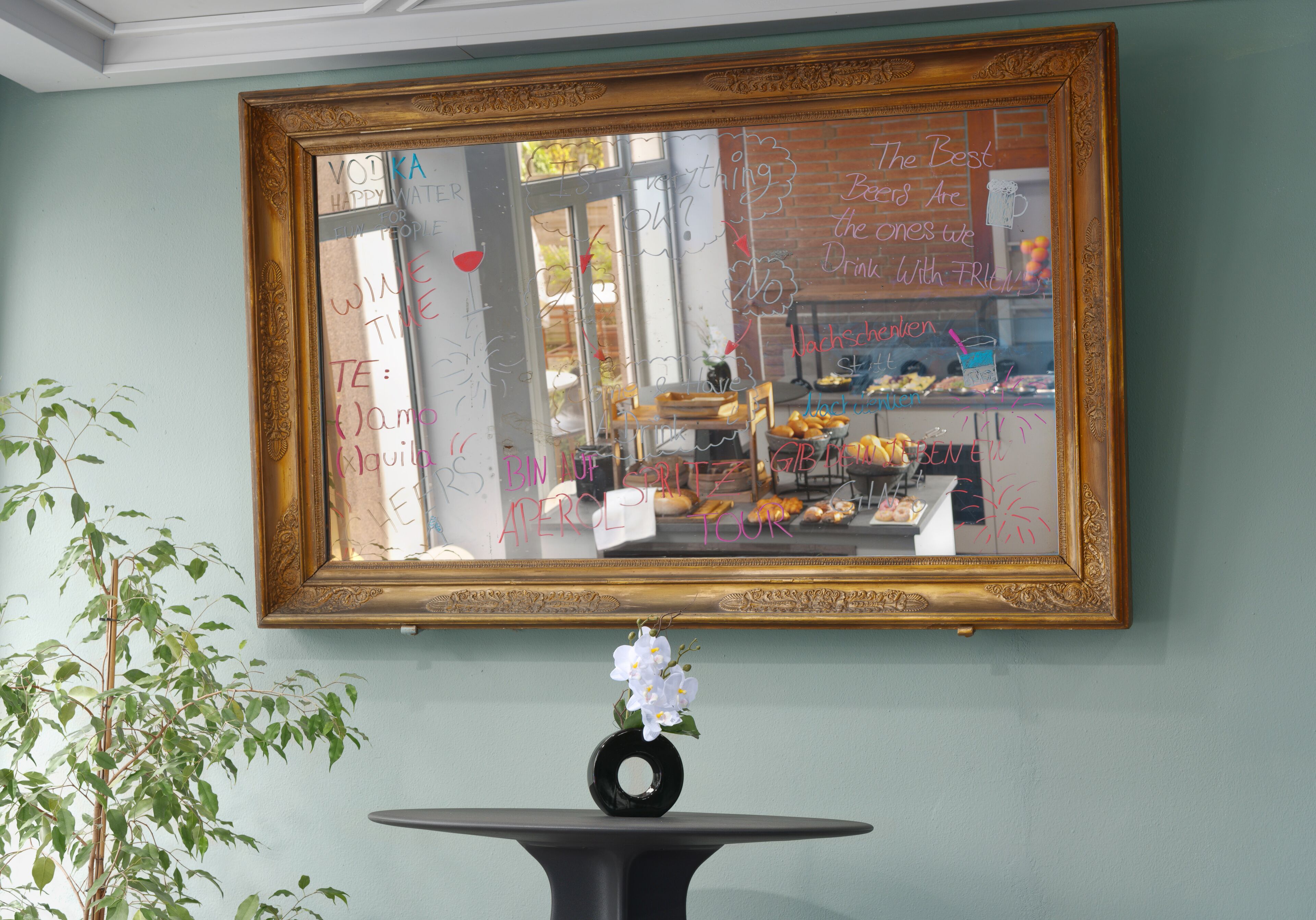 Espejo enmarco que refleja el interior de una panadería, con una mesa negra y un jarrón con flores blancas al frente.
