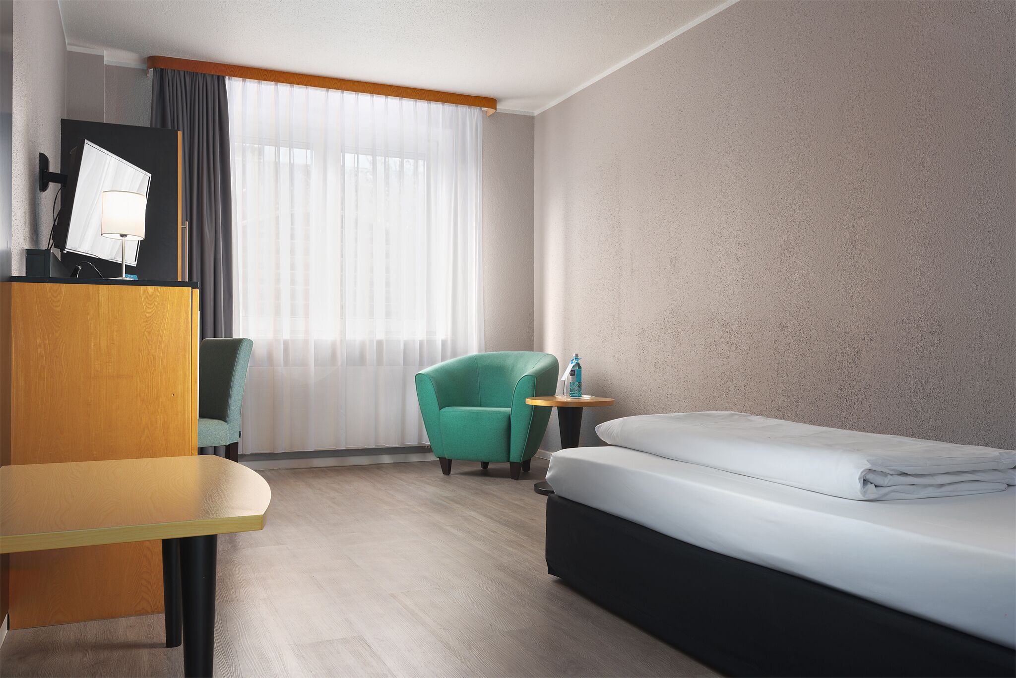 A hotel room with a bed, a desk, a chair, and a small table with a bottle.
