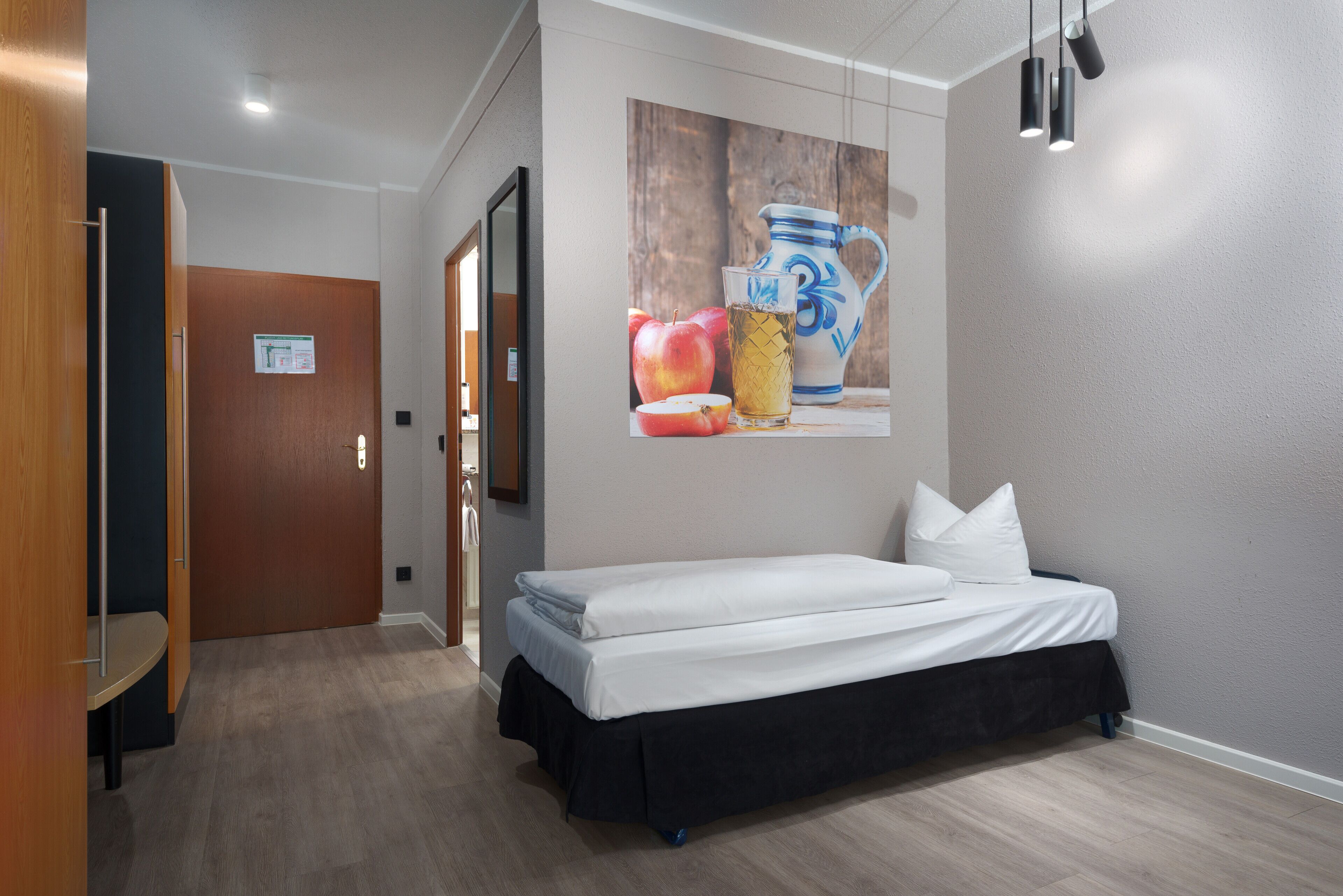 A hotel room with a bed, a wooden door, a wall-mounted picture, and a light fixture.