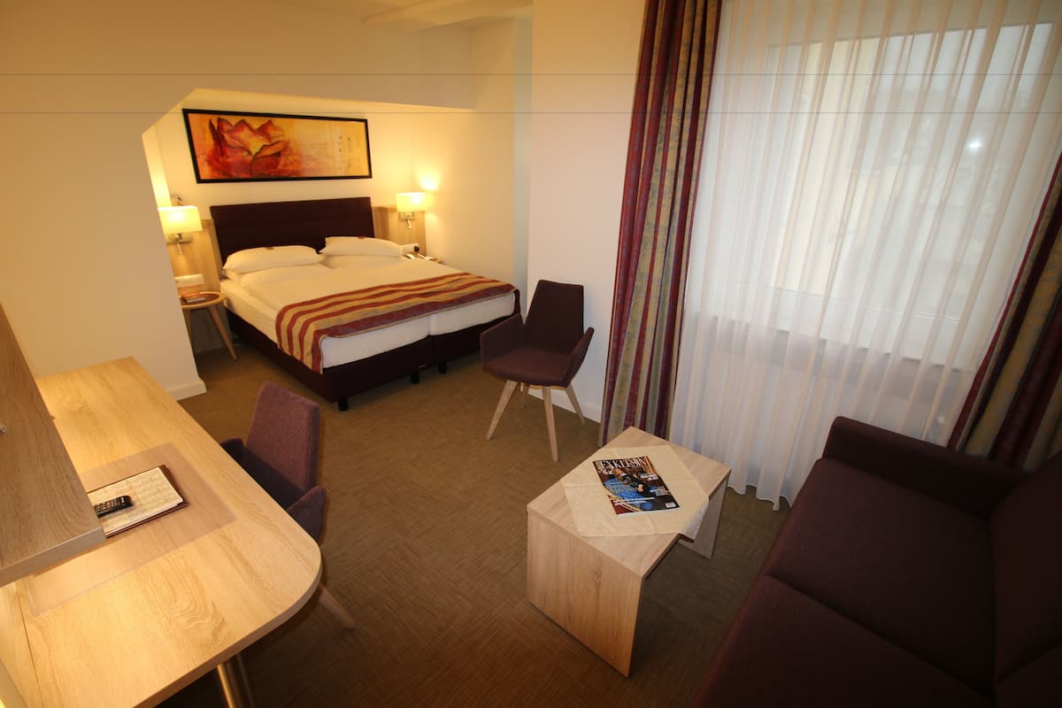 A hotel room with a bed, a desk, a chair, a small table, and a window with curtains.