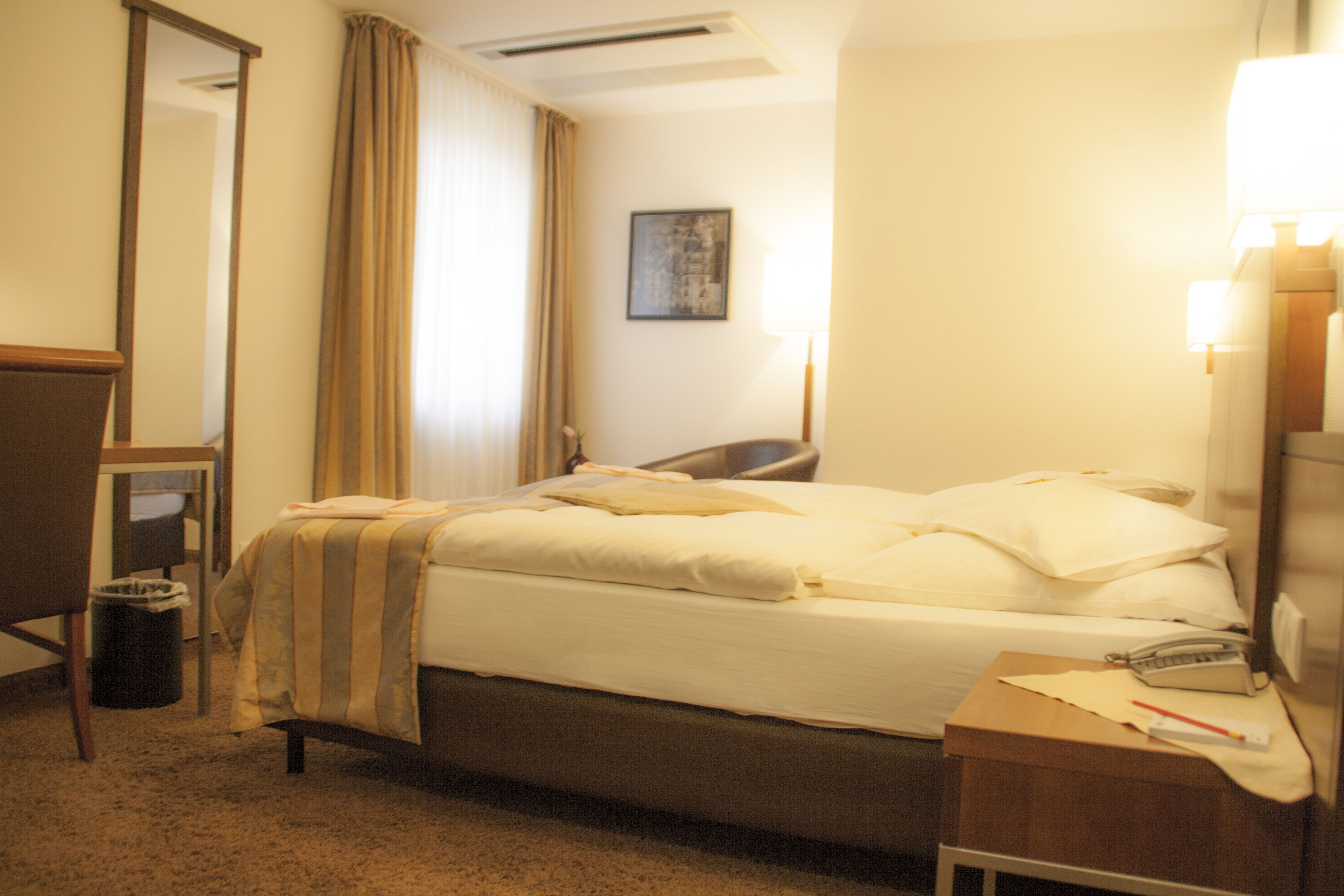 Une chambre d’hôtel avec un lit, un bureau, une chaise, une poubelle et une lampe.