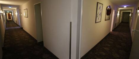 A hotel hallway with carpeted floors, doors, and framed artwork on the walls.