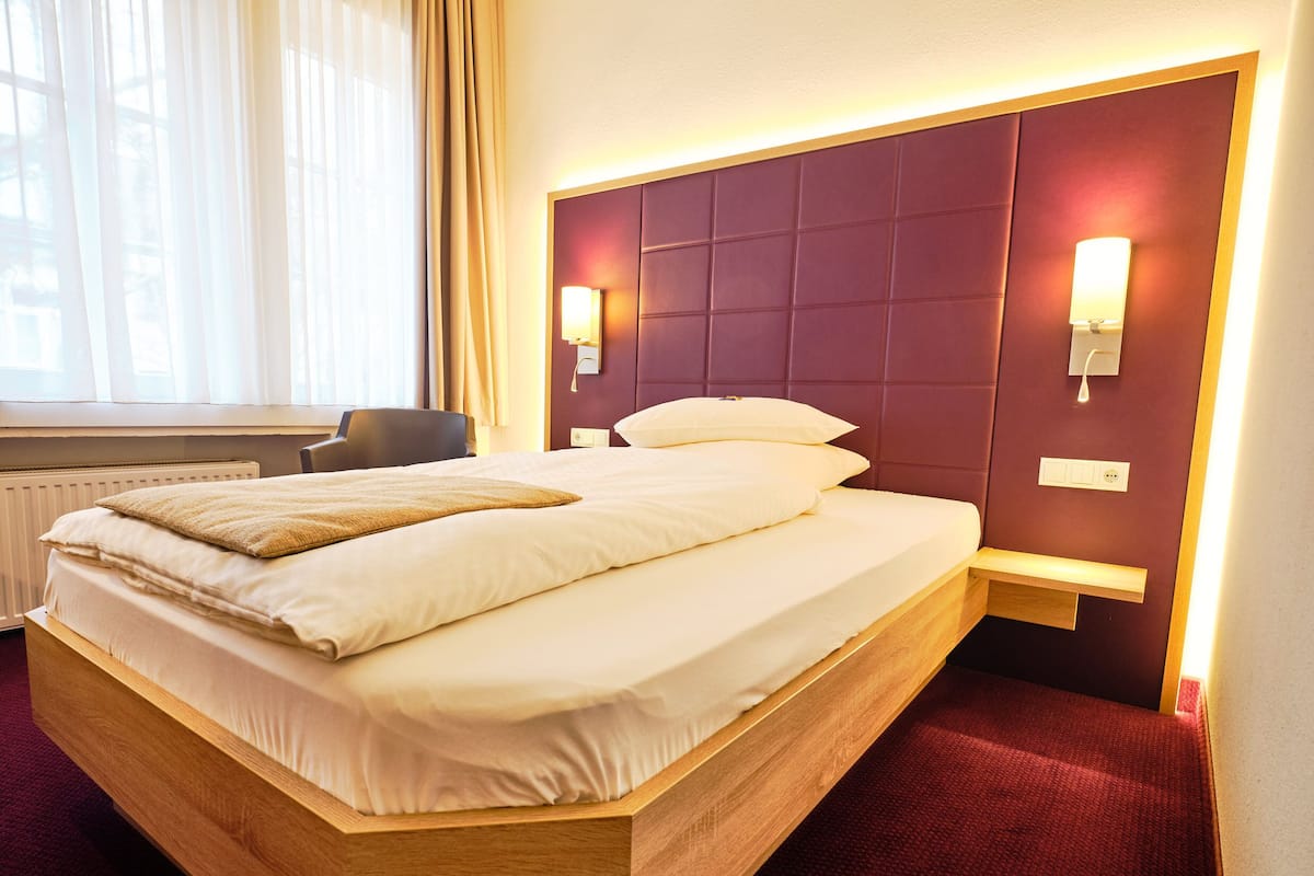 A neatly made bed with a wooden headboard, a bedside table with a lamp, and a chair in a hotel room.