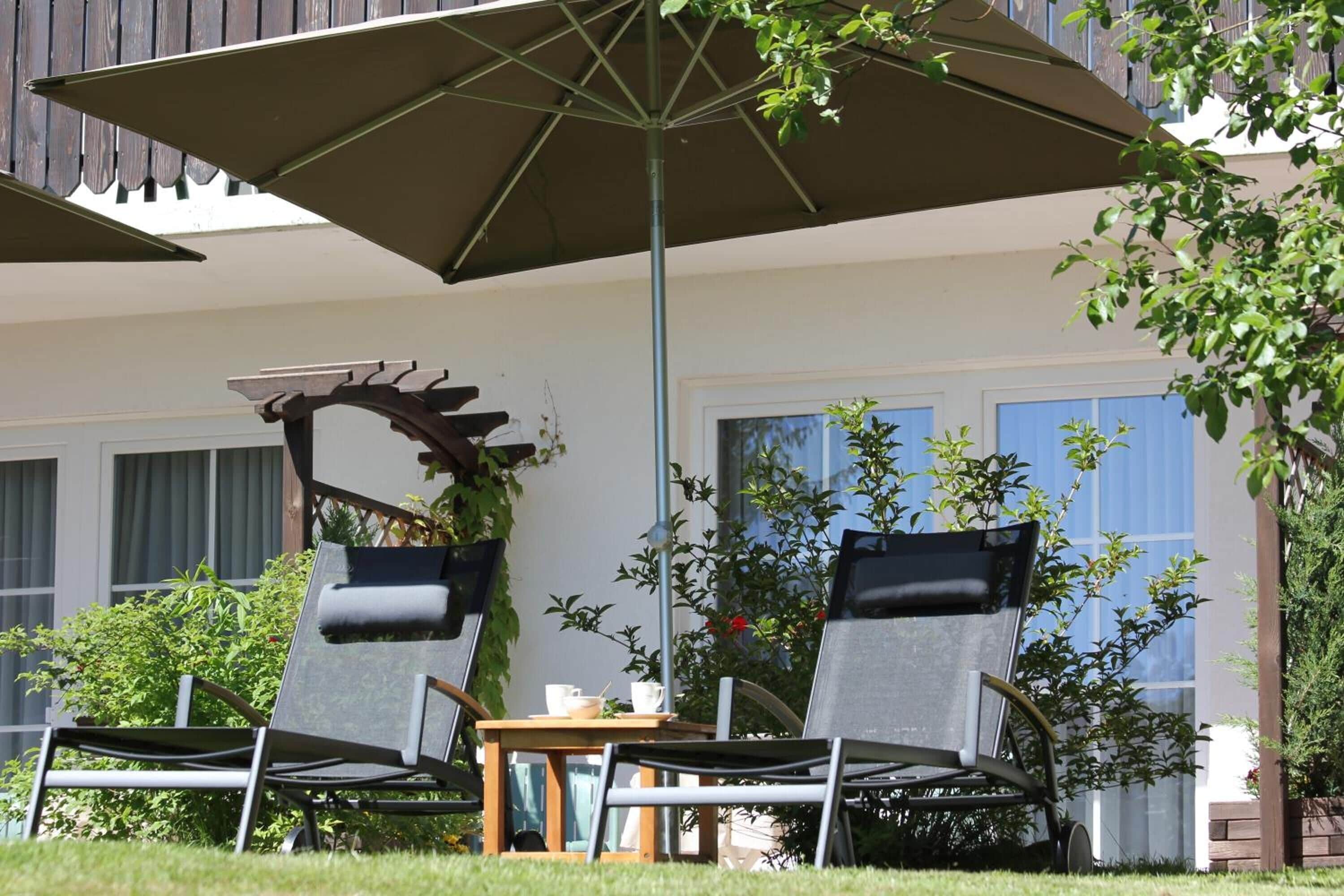 Une terrasse avec deux chaises et une table, protégée par un grand parasol, entourée de verdure et bordée d’un bâtiment doté de fenêtres.