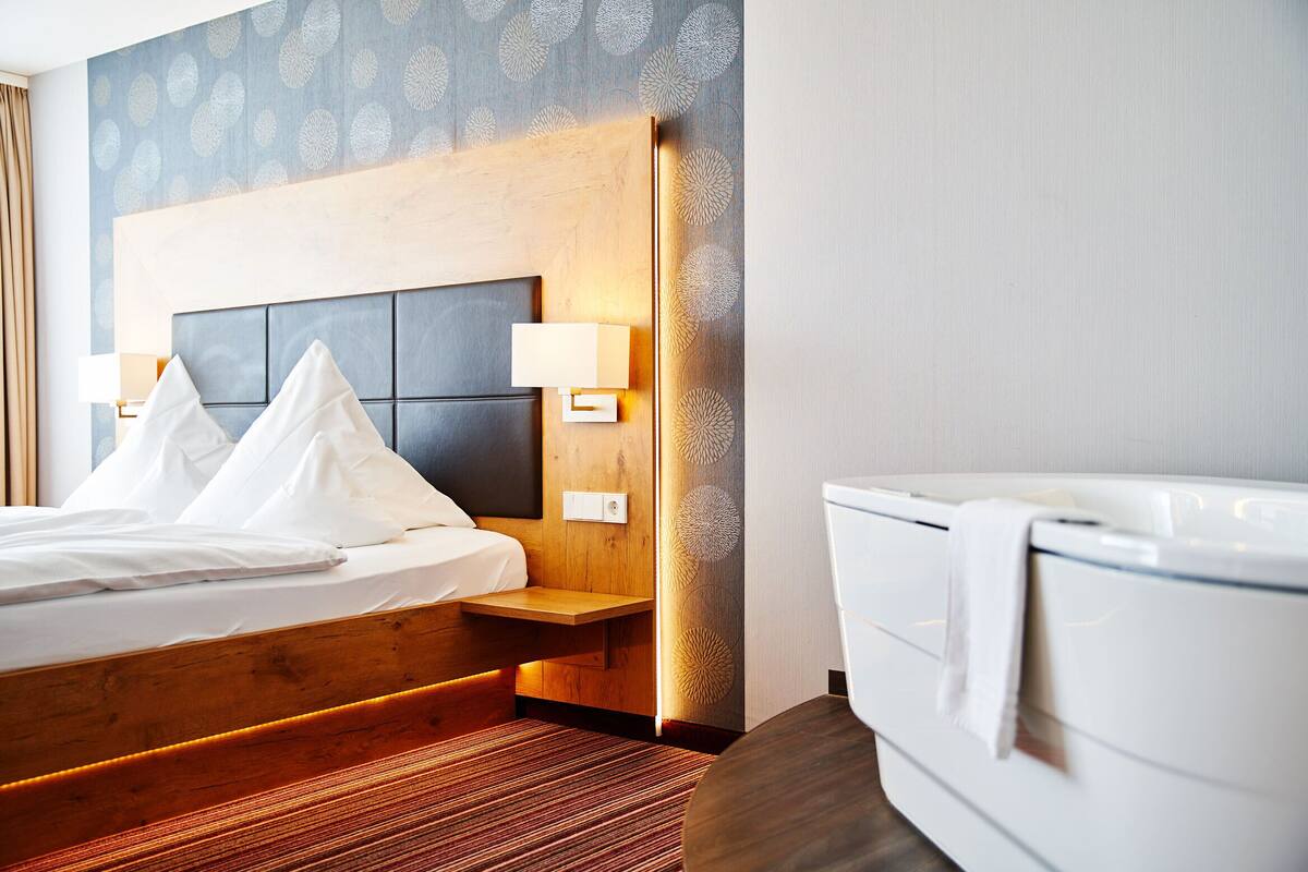 A hotel room with a wooden headboard, a bed with white linens, a bedside table with a lamp, and a white bathtub.
