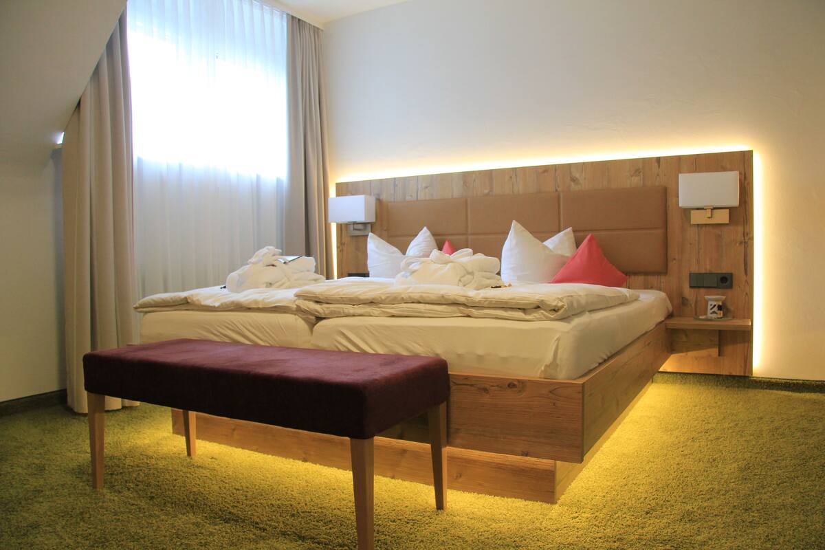 A hotel room with a large bed, a wooden headboard, a bench, and a nightstand with a lamp.