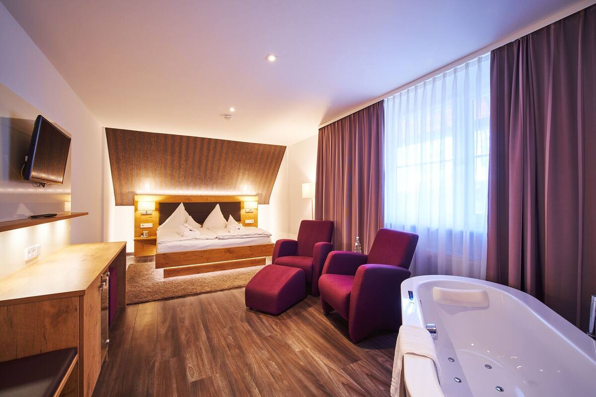 A modern hotel room with a large bed, a sitting area with armchairs, a desk, a TV, and a bathtub.