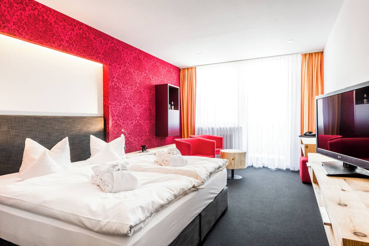 A modern hotel room with a large bed, a TV, a red sofa, and a wooden side table.