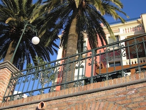 A brick wall with a metal railing, palm trees, and a building with balconies. - Hotel Villa Schuler (Taormina)