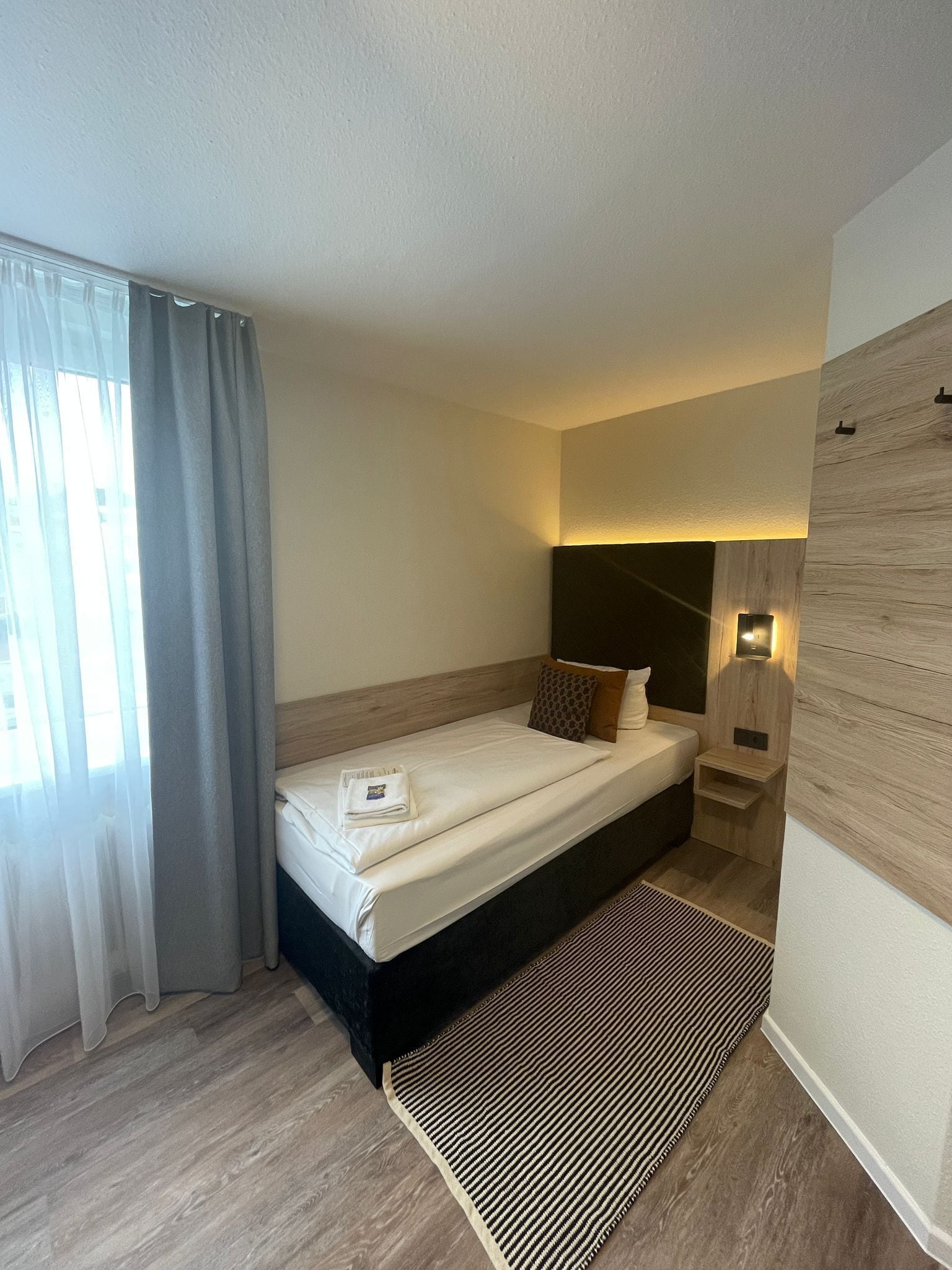 Habitación de hotel moderna con una cama individual, una mesita de noche, una lámpara de pared y un ventanal con cortinas.