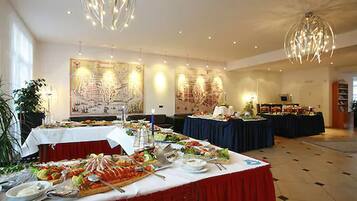 A buffet setup with multiple tables, each covered with a white tablecloth. There are various food items and utensils on the tables. The room has a high ceiling with hanging light fixtures and large windows allowing natural light.