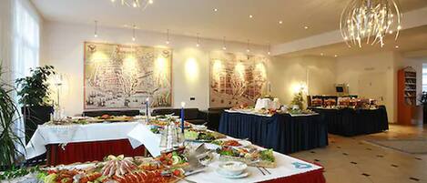 A buffet setup with multiple tables, each covered with a white tablecloth. There are various food items and utensils on the tables. The room has a high ceiling with hanging light fixtures and large windows allowing natural light.