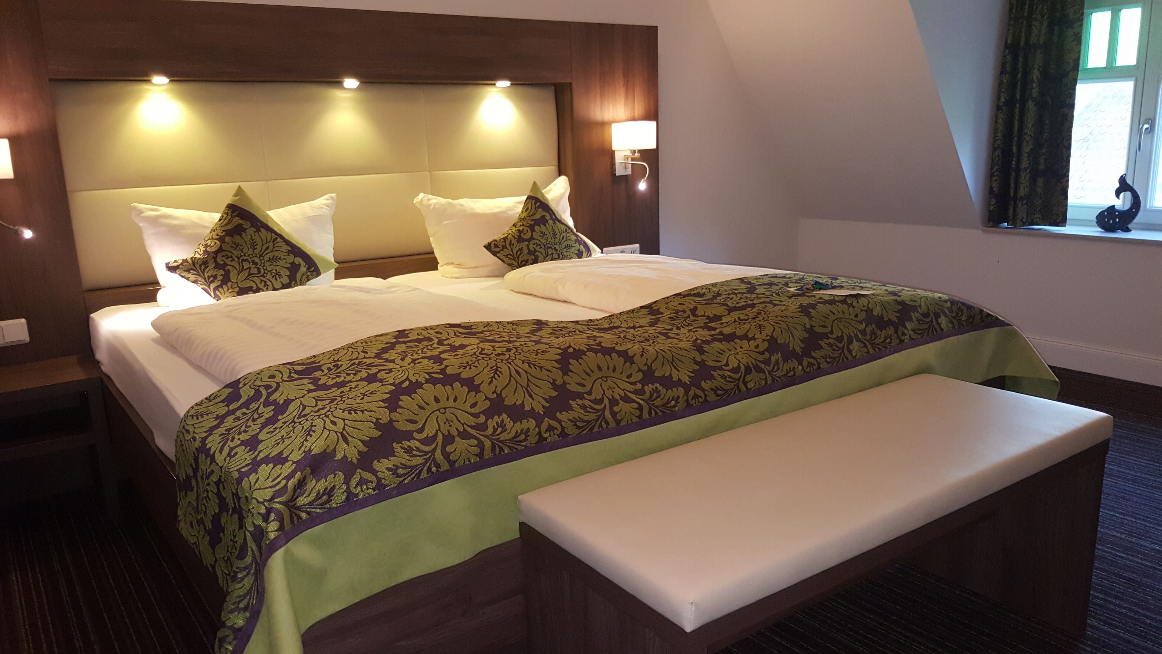 A hotel room with a large bed, a bench, a headboard with lights, and a window with curtains.