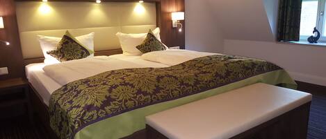 A hotel room with a large bed, a bench, a headboard with lights, and a window with curtains.