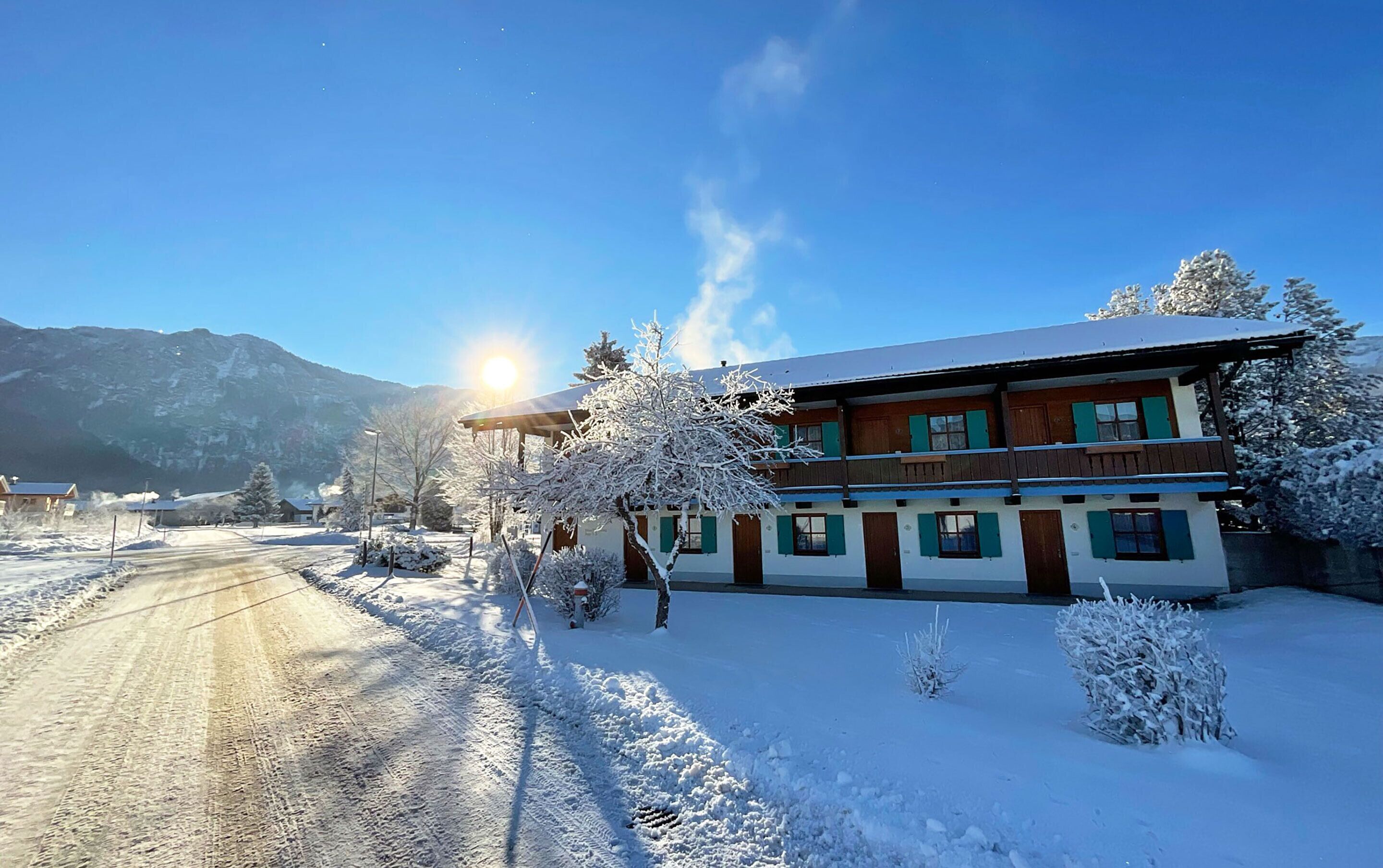 A two-story building with a snow-covered roof and a balcony. There is a clear sky and mountains in the background.