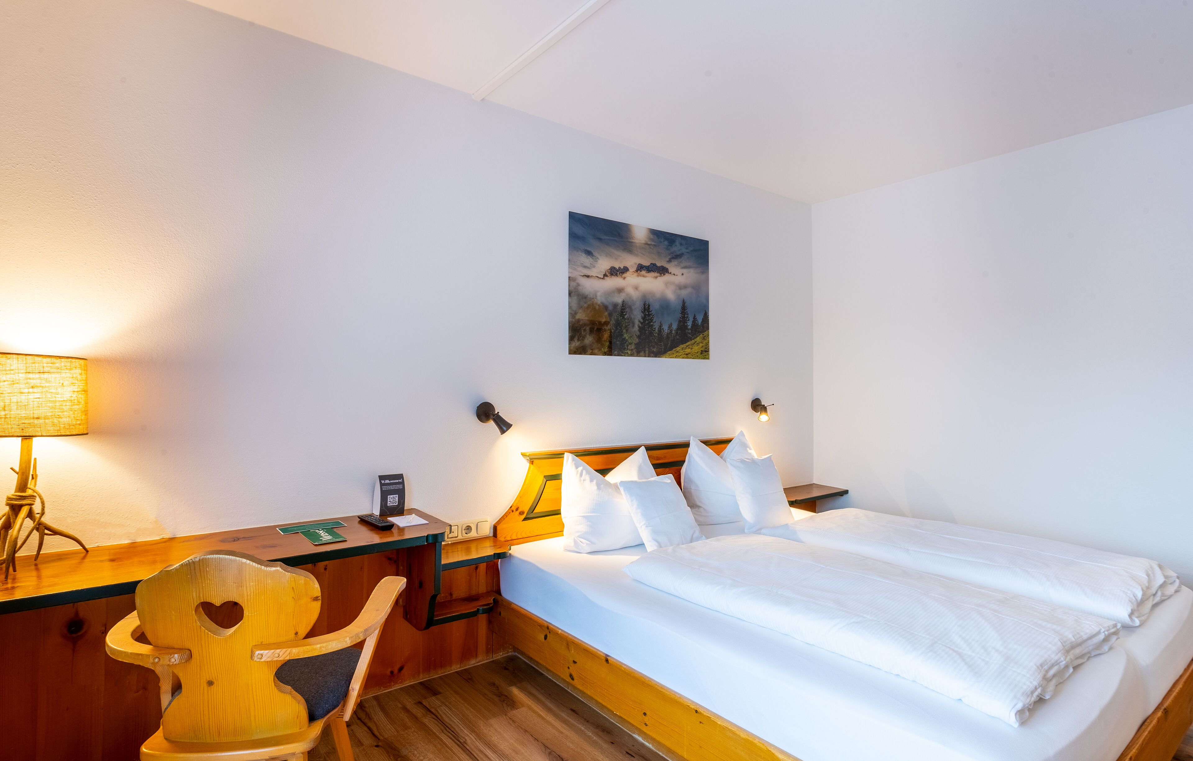 Ein Hotelzimmer mit einem hölzernen Bett, einem Schreibtisch mit Lampe, einem Sessel und einem Bild an der Wand.