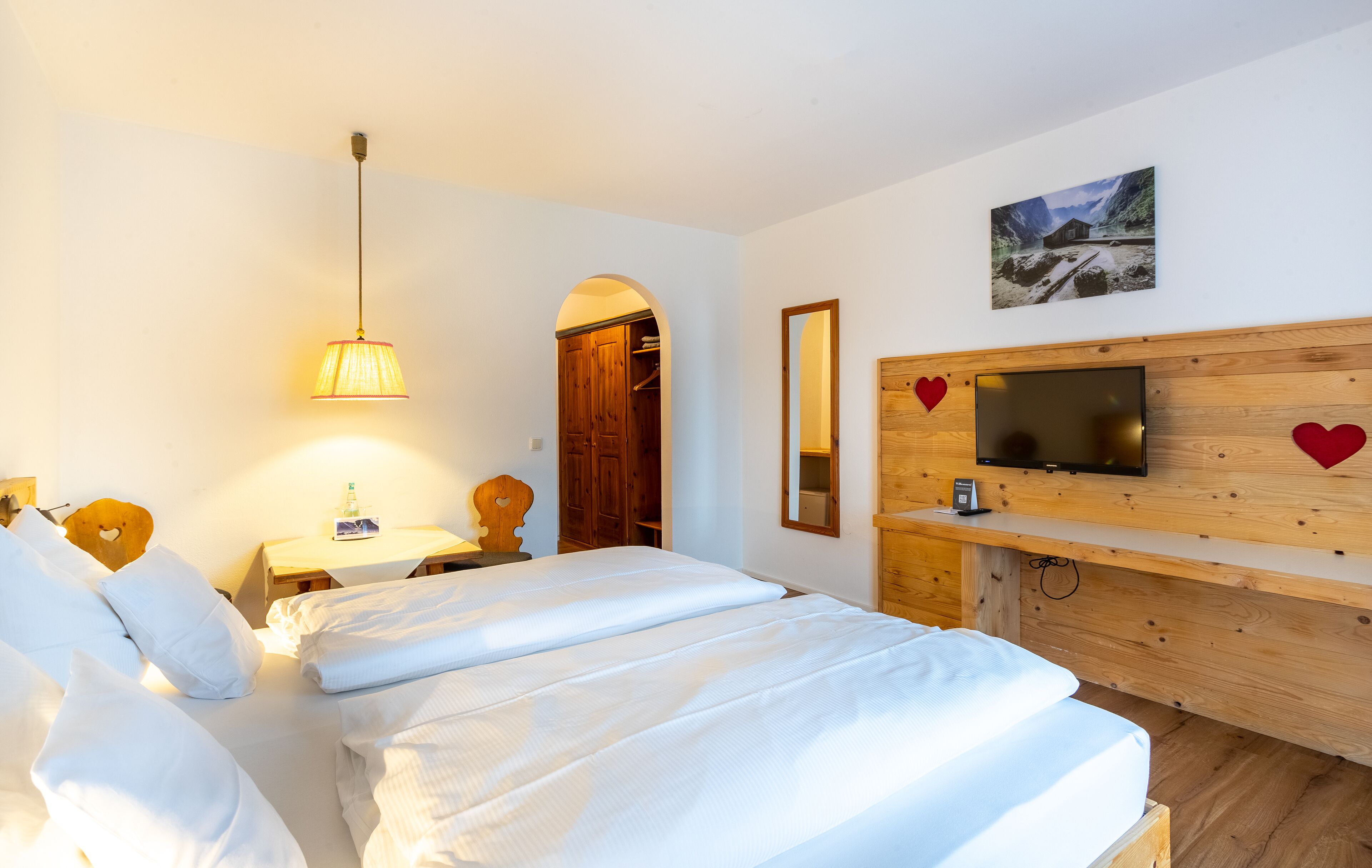 A hotel room with a bed, a TV mounted on a wooden wall, a wooden wardrobe, and a small table with a lamp.
