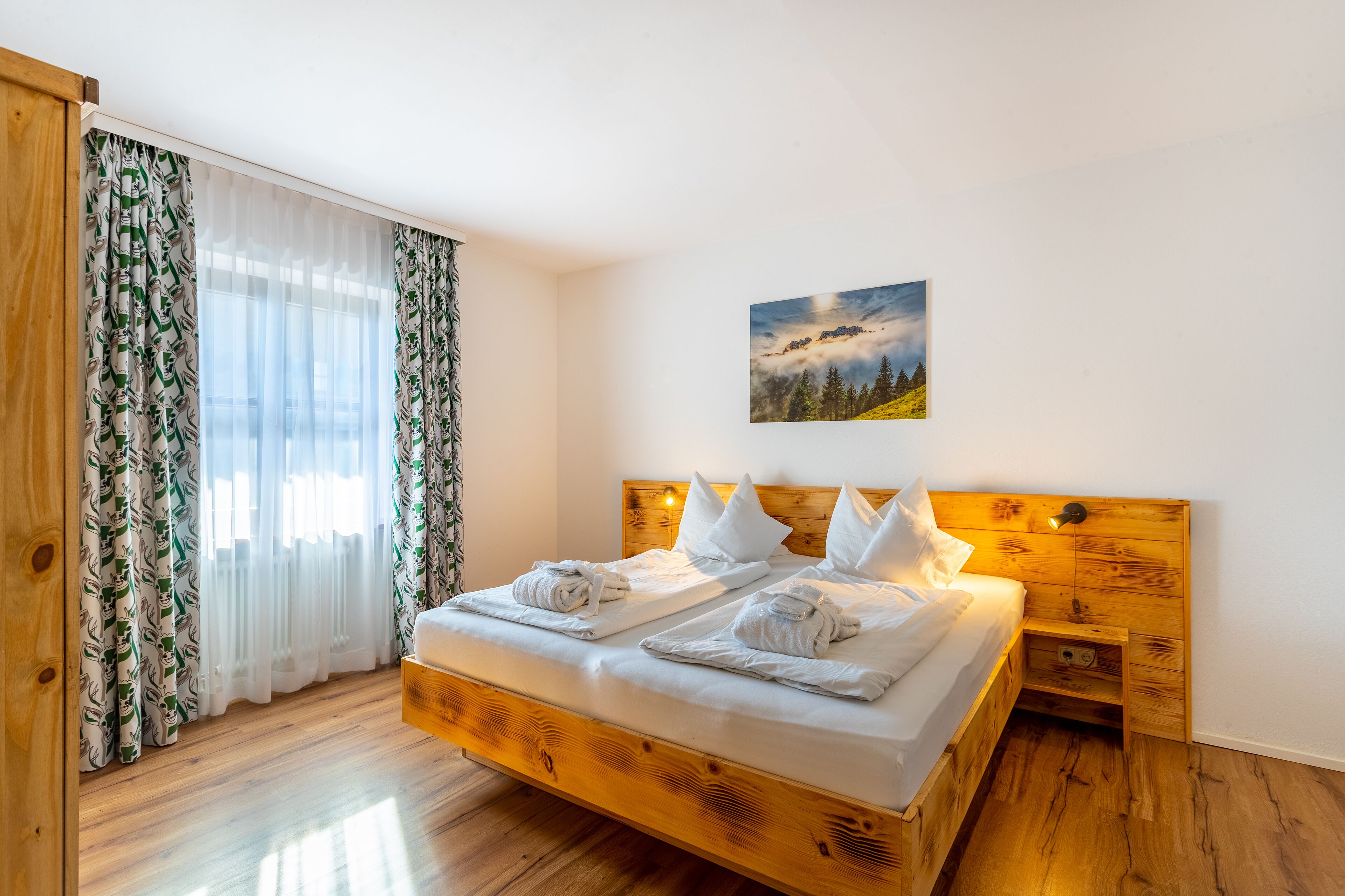 Ein Bett aus Holz mit weißen Leinen und Handtüchern, ein Nachttisch, ein Gemälde mit Berglandschaft und leichte Vorhänge mit Blätermotiv.